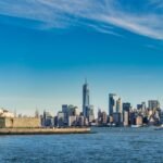 Statue of Liberty on Liberty Island with the Manhattan skyline across the harbor behind her, under a blue sky.