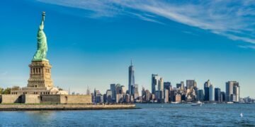 Statue of Liberty on Liberty Island with the Manhattan skyline across the harbor behind her, under a blue sky.