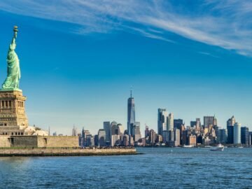 Statue of Liberty on Liberty Island with the Manhattan skyline across the harbor behind her, under a blue sky.