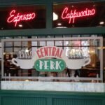 Neon red 'Espresso' and 'Cappuccino' signs in a cafe window above a Central Perk logo.