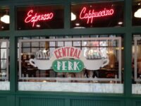 Neon red 'Espresso' and 'Cappuccino' signs in a cafe window above a Central Perk logo.